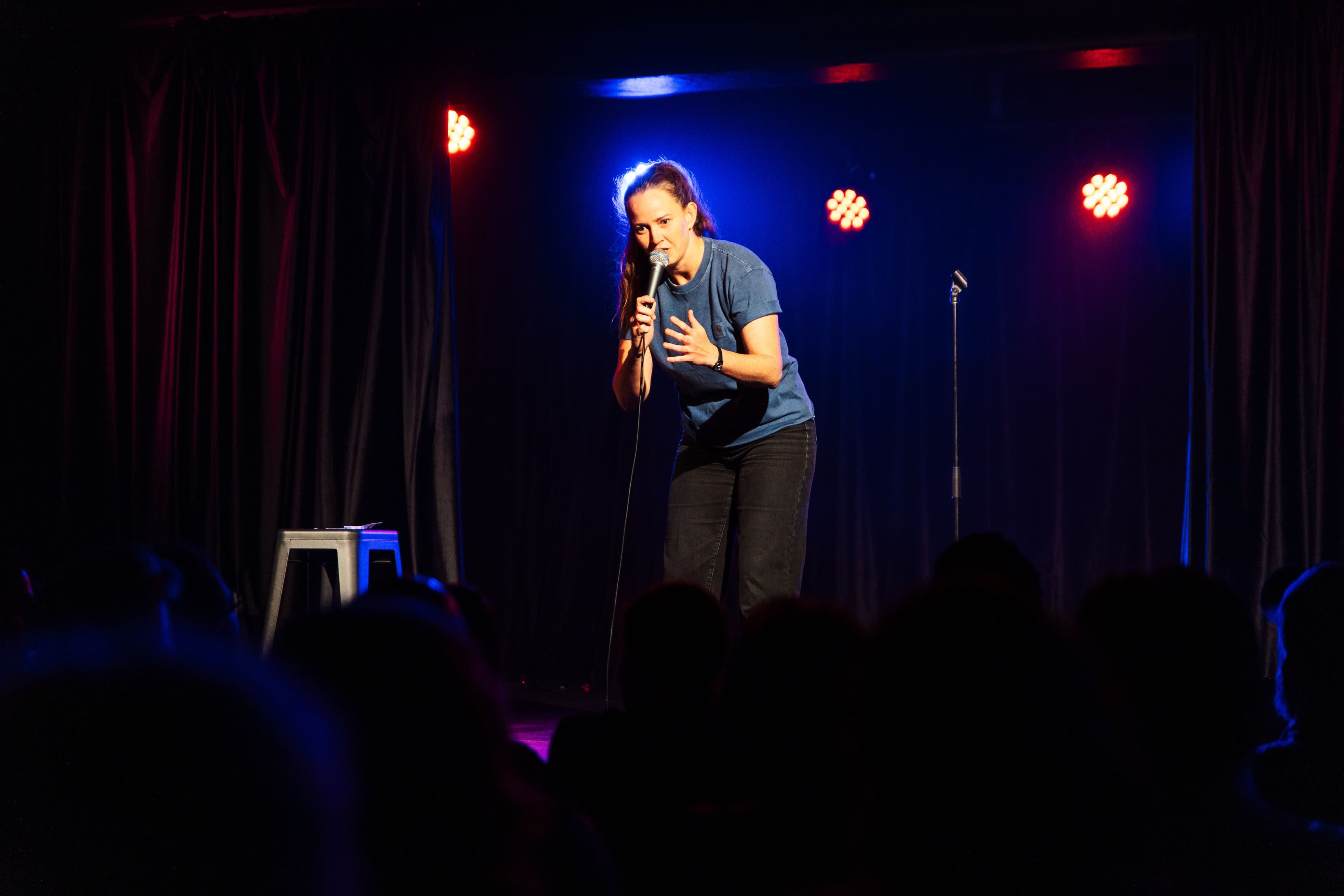 A comedian in a blue shirt performs on stage with a microphone, engaging the audience under red and blue lights.